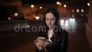 午夜美丽的女孩使用智能手机午夜女孩站在街道上，靠近道路，使用智能手机